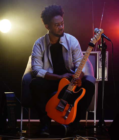 Zeruabbel Molla sitting on stage holding an electric guitar with stage lights in the background.