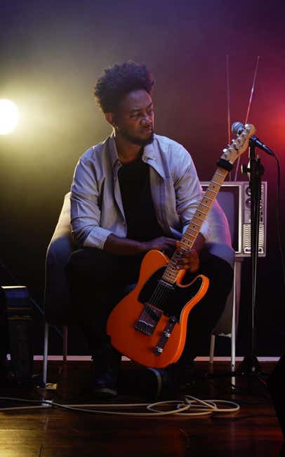Zeruabbel Molla sitting on a chair holding an electric guitar on stage with colorful lights.