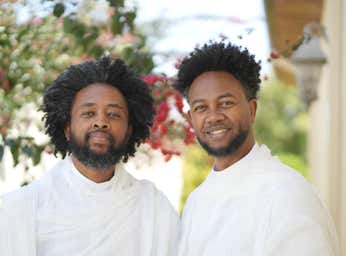 Zeruabbel Molla and Yamlu Molla wearing white clothing, standing outdoors with greenery and flowers in the background.